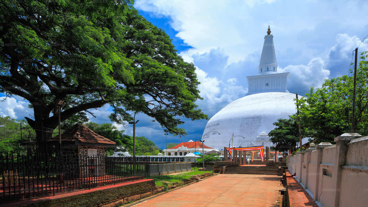 A view of the Sacred City of Anuradhapura with its ancient ruins, including a large white temple and a smaller black structure, surrounded by green trees and a blue sky with clouds.