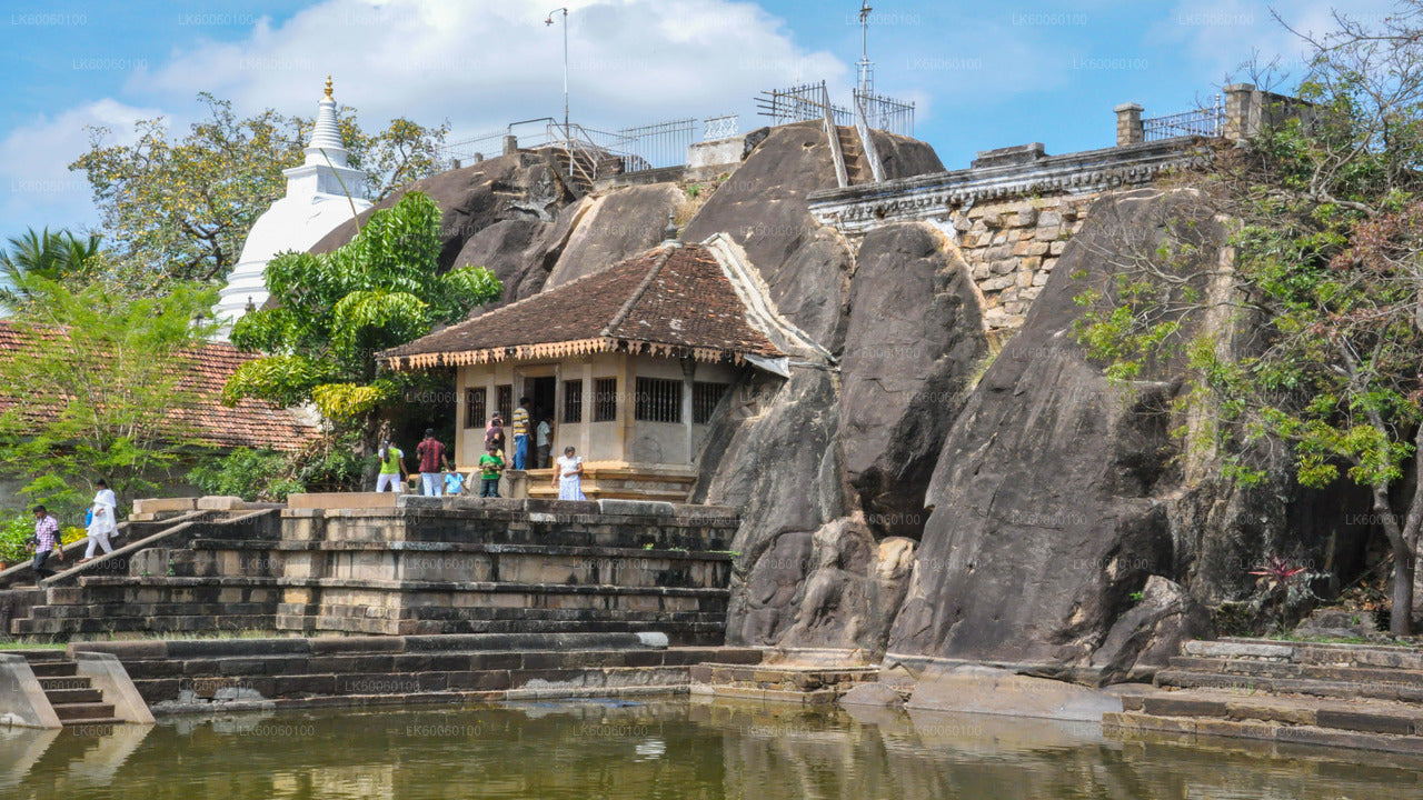 Ancient rock temple complex with a white stupa, shrine, and stone steps beside a pond, surrounded by large boulders.